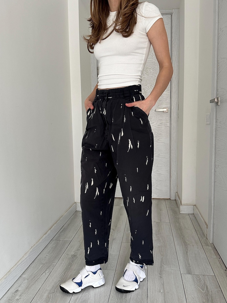 Puede incluir: Pantalones negros con trazos de pintura abstractos blancos. Los pantalones tienen cintura alta y corte holgado. La persona lleva una camiseta blanca y zapatillas blancas y azules.