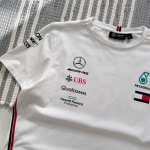 May include: White t-shirt with the Mercedes-AMG Petronas Formula One Team logo and sponsors including UBS, Qualcomm, and Hewlett Packard Enterprise.