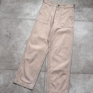 Puede incluir: Pantalones color beige con dise&ntilde;o de pernera ancha. Los pantalones tienen dos bolsillos delanteros grandes y cierre de bot&oacute;n. La tela parece ser de algod&oacute;n resistente, adecuada para uso casual. Los pantalones est&aacute;n planos sobre una superficie gris.