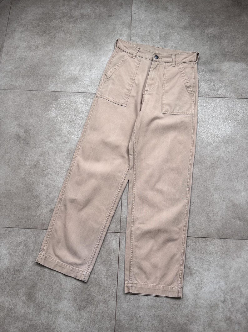 Puede incluir: Pantalones color beige con dise&ntilde;o de pernera ancha. Los pantalones tienen dos bolsillos delanteros grandes y cierre de bot&oacute;n. La tela parece ser de algod&oacute;n resistente, adecuada para uso casual. Los pantalones est&aacute;n planos sobre una superficie gris.