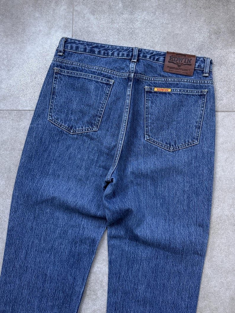 Pode incluir: Cal&ccedil;as de ganga azuis com dois bolsos traseiros e um remendo de couro castanho com a marca "EDWIN". As cal&ccedil;as de ganga t&ecirc;m um design cl&aacute;ssico de cinco bolsos e um corte reto. A ganga tem uma lavagem m&eacute;dia.