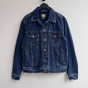 Puede incluir: Chaqueta vaquera azul oscuro con un diseño clásico. Incluye cierre de botones, dos bolsillos en el pecho y dos bolsillos laterales. La chaqueta tiene cuello y está colgada de una percha negra.