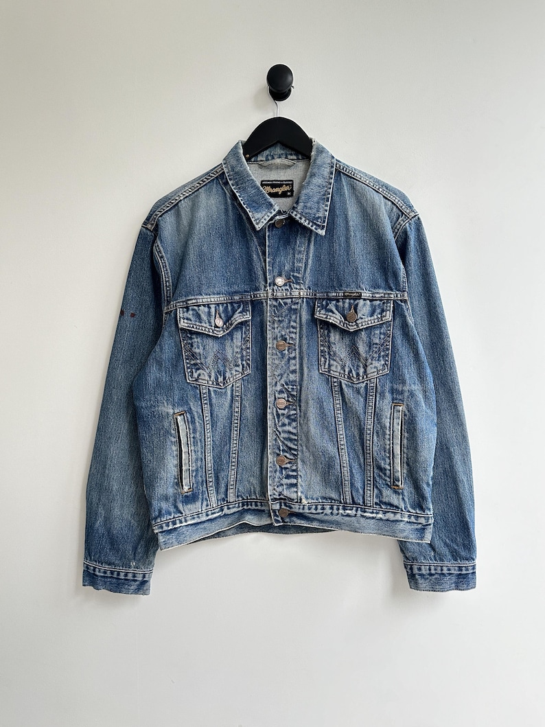 May include: A blue denim jacket with a Wrangler logo on the pocket. The jacket has a button closure and two pockets on the chest.