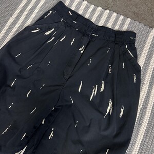 Puede incluir: Pantalones azul marino oscuro con cintura alta y estampado de pinceladas blancas. Los pantalones tienen pliegues en la cintura y bolsillos. La tela parece ser de peso medio.