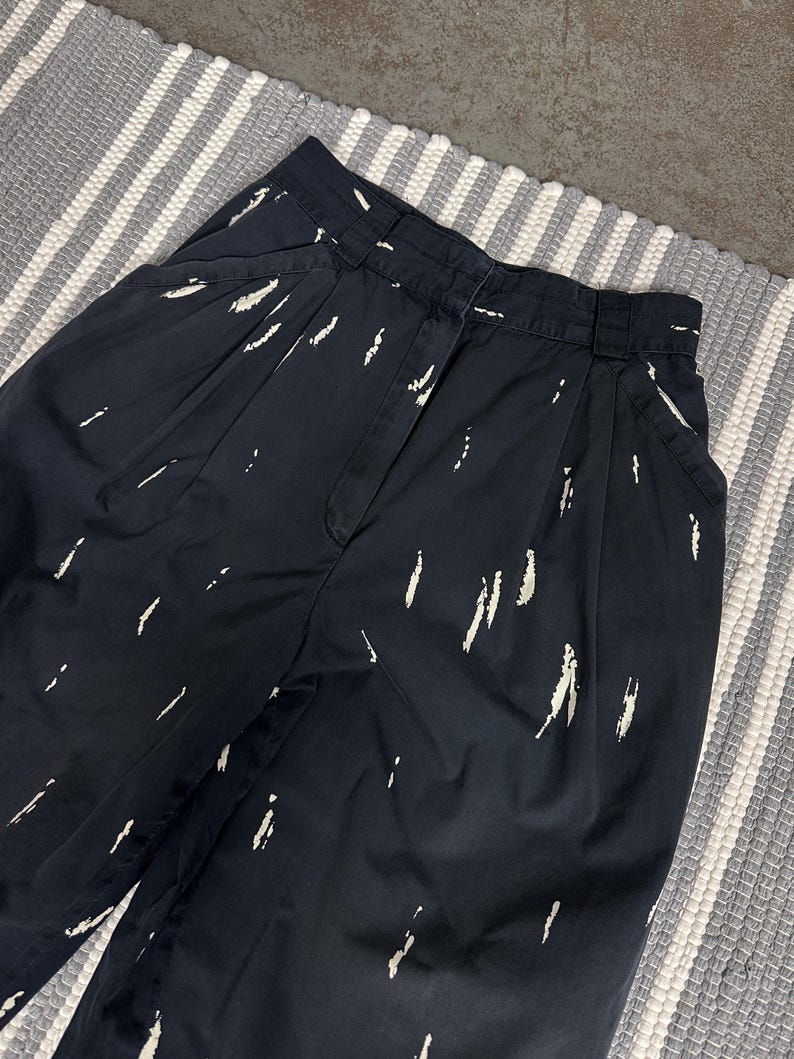Puede incluir: Pantalones azul marino oscuro con cintura alta y estampado de pinceladas blancas. Los pantalones tienen pliegues en la cintura y bolsillos. La tela parece ser de peso medio.