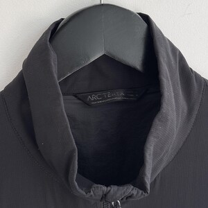 May include: Black jacket with a high collar and a full-length zipper. The jacket is hanging on a black hanger. The brand name "Arc'teryx" is visible on the label.