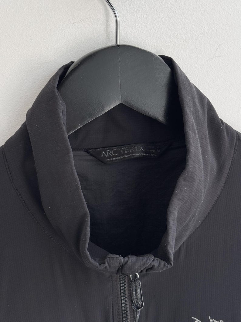 May include: Black jacket with a high collar and a full-length zipper. The jacket is hanging on a black hanger. The brand name "Arc'teryx" is visible on the label.