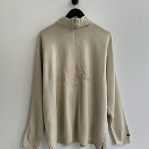 Puede incluir: Jersey de punto beige con cuello alto y cierre de cremallera de cuarto. El jersey presenta un diseño bordado en el pecho y un pequeño logotipo en el puño. El jersey de manga larga está colgado de una percha negra.