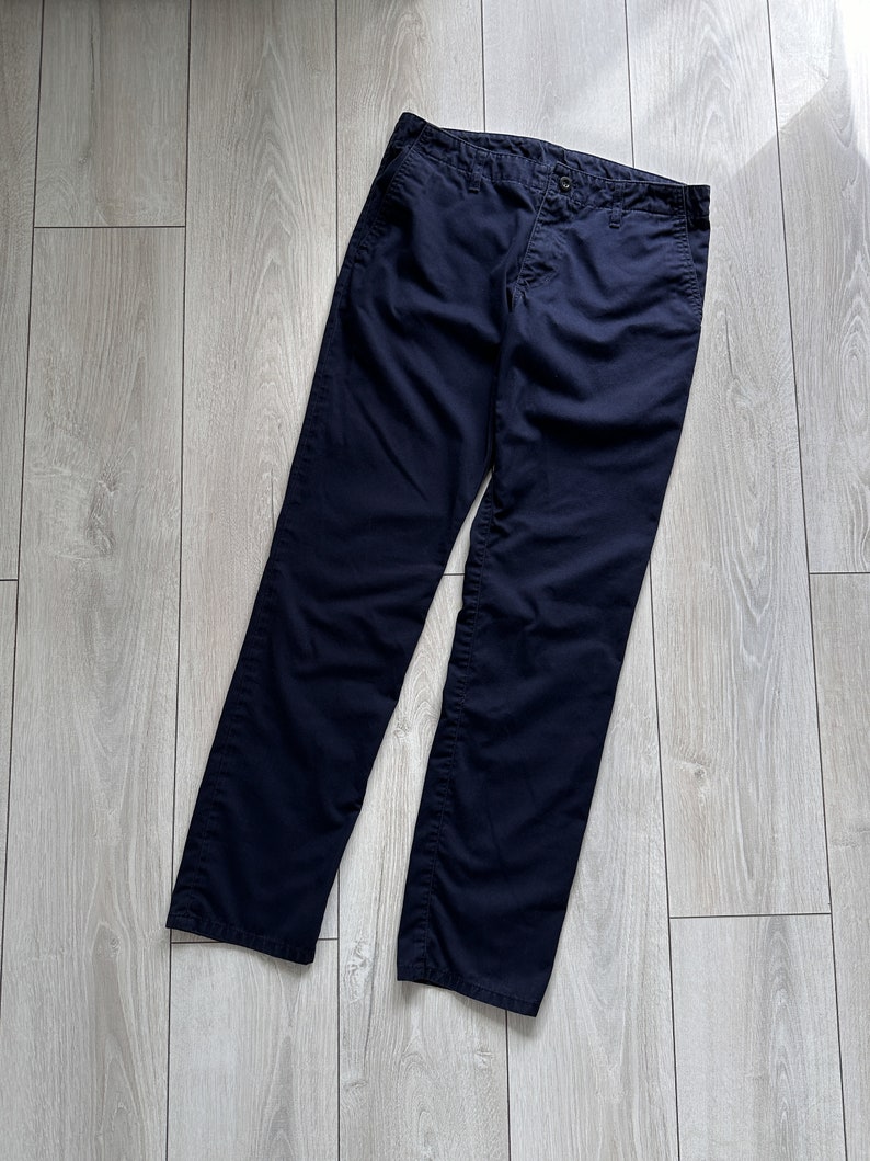 K&ouml;nnte beinhalten: Marineblaue Hose aus strapazierf&auml;higem Stoff. Die Hose hat einen Knopfverschluss und ein klassisches gerades Bein-Design. Das Kleidungsst&uuml;ck liegt flach auf einer hellen Holzoberfl&auml;che.