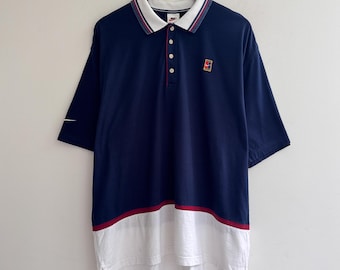 Könnte beinhalten: Ein dunkelblaues Poloshirt mit einem weißen unteren Bereich, der durch einen dünnen roten Streifen abgesetzt ist. Das Shirt hat einen weißen Kragen mit dunkelblau-rotem Besatz, eine Knopfleiste mit drei Knöpfen, ein Nike-Tennislogo und einen weißen Swoosh.