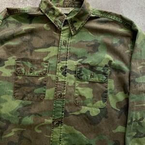 May include: A green, brown, and black camouflage long-sleeve button-up shirt with two chest pockets. The shirt has a label that reads "Dave Frohtier" on the left side of the collar.