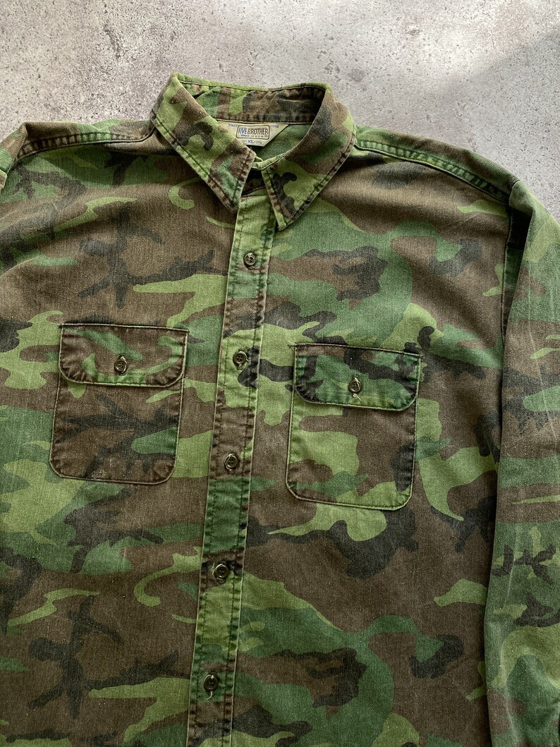 May include: A green, brown, and black camouflage long-sleeve button-up shirt with two chest pockets. The shirt has a label that reads "Dave Frohtier" on the left side of the collar.
