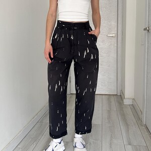 Puede incluir: Pantalones negros con patrones blancos en forma de pinceladas. Los pantalones de cintura alta tienen un corte holgado. La persona lleva una camiseta blanca y zapatillas blancas con detalles azules. El atuendo es casual y elegante.