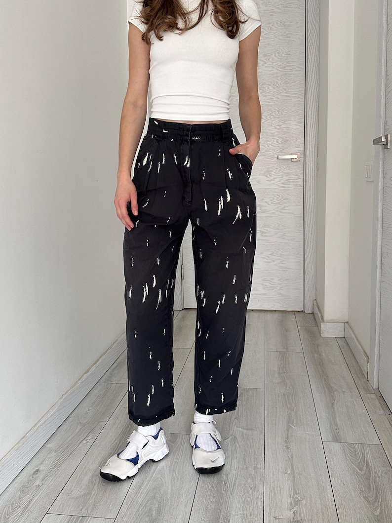 Puede incluir: Pantalones negros con patrones blancos en forma de pinceladas. Los pantalones de cintura alta tienen un corte holgado. La persona lleva una camiseta blanca y zapatillas blancas con detalles azules. El atuendo es casual y elegante.