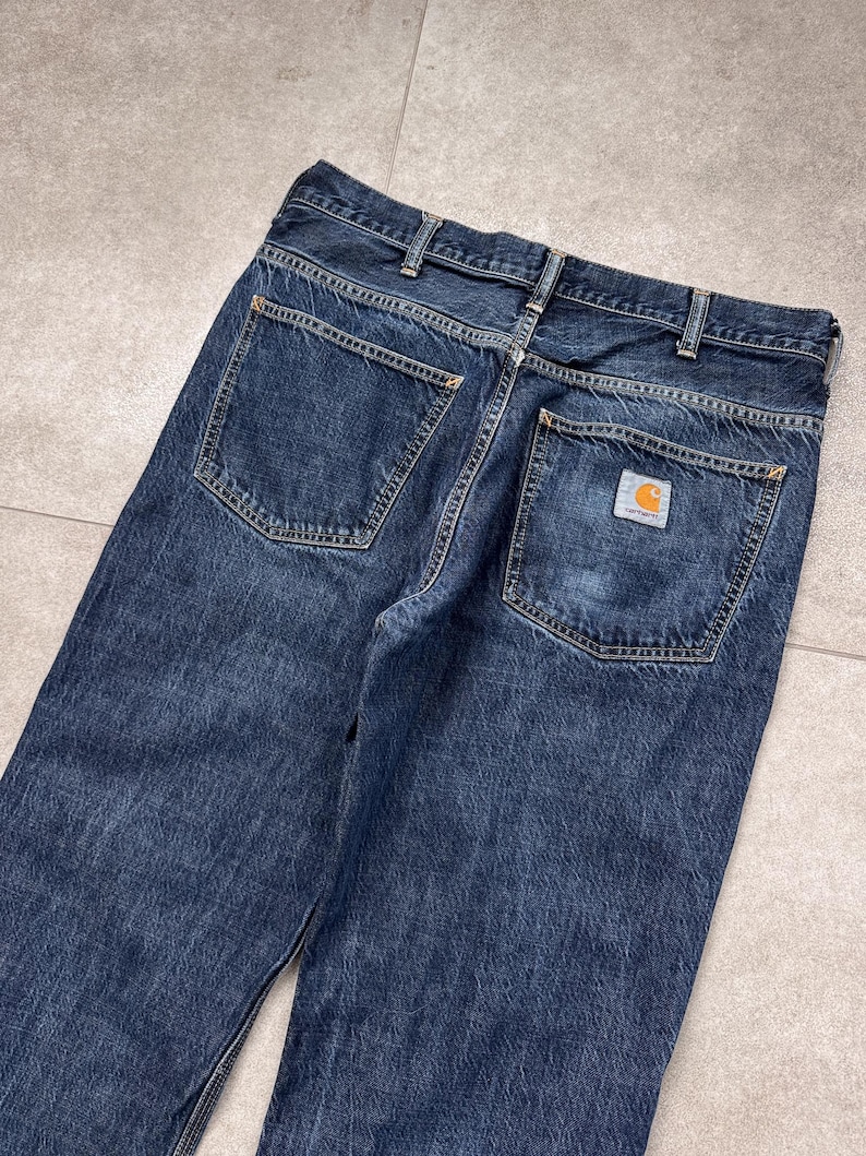 Puede incluir: Vaqueros de mezclilla azul oscuro con dos bolsillos traseros y un peque&ntilde;o parche cuadrado con el logotipo. Los vaqueros tienen un dise&ntilde;o cl&aacute;sico de cinco bolsillos y un corte recto. Las costuras son de color dorado en contraste.