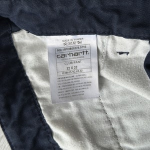 K&ouml;nnte beinhalten: Nahaufnahme einer marineblauen Carhartt Club Hose. Das Etikett lautet "32 x 32" (81 x 81 cm). Das Etikett enth&auml;lt auch den Text "MADE IN TUNISIA" und "WORK IN PROGRESS". Die Hose besteht aus einer Baumwoll- und Polyestermischung.