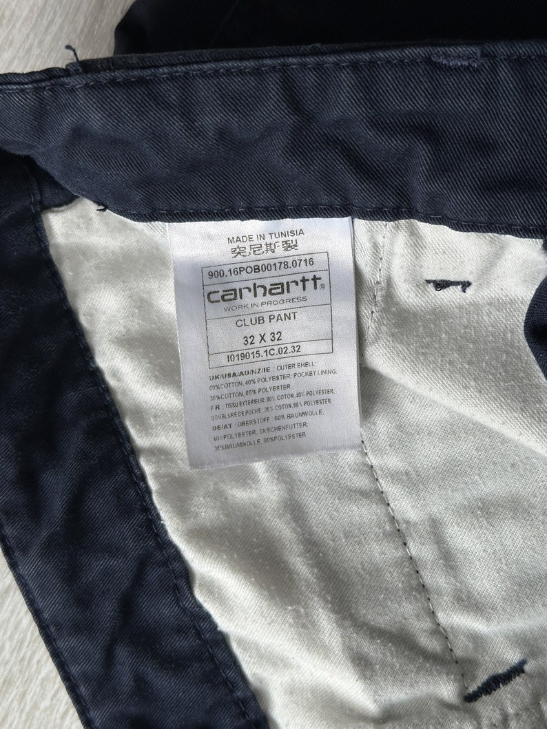 K&ouml;nnte beinhalten: Nahaufnahme einer marineblauen Carhartt Club Hose. Das Etikett lautet "32 x 32" (81 x 81 cm). Das Etikett enth&auml;lt auch den Text "MADE IN TUNISIA" und "WORK IN PROGRESS". Die Hose besteht aus einer Baumwoll- und Polyestermischung.