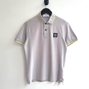 Pode incluir: Uma camisa polo roxa clara com acabamento amarelo e branco na gola e nas mangas. A camisa tem um pequeno patch quadrado no peito e uma carcela de dois botões. Está pendurada num cabide preto.