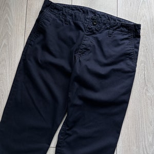 K&ouml;nnte beinhalten: Dunkelmarineblaue Hose mit Knopfverschluss und Taschen. Die Hose ist aus einem einfarbigen, dunkelblauen Stoff gefertigt. Das Kleidungsst&uuml;ck liegt flach auf einer hellen Holzmaserung.