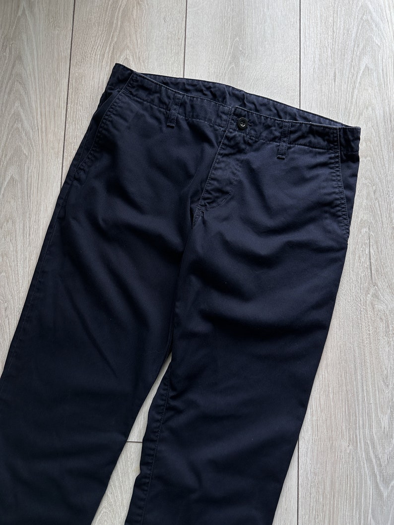 K&ouml;nnte beinhalten: Dunkelmarineblaue Hose mit Knopfverschluss und Taschen. Die Hose ist aus einem einfarbigen, dunkelblauen Stoff gefertigt. Das Kleidungsst&uuml;ck liegt flach auf einer hellen Holzmaserung.