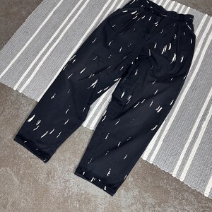 Puede incluir: Pantalones negros con un patr&oacute;n de pinceladas blancas. Los pantalones tienen la parte delantera plisada y un corte holgado. Se encuentran sobre una alfombra a rayas grises y blancas. La tela parece ligera.