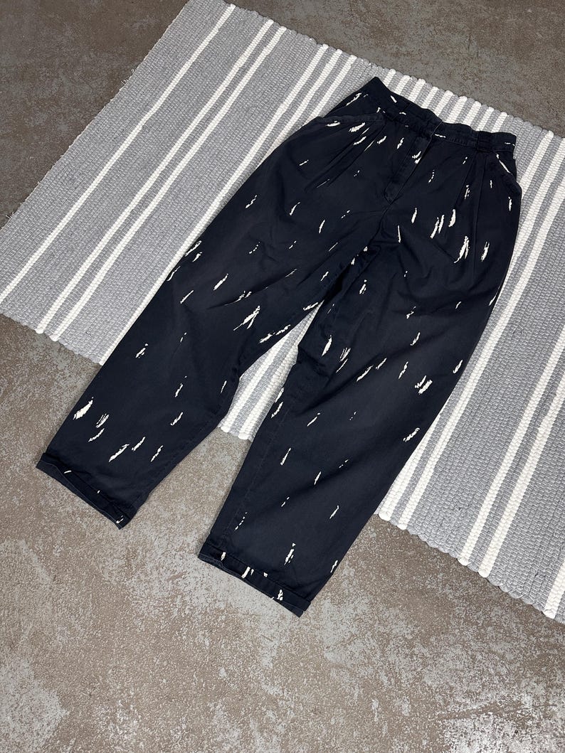 Puede incluir: Pantalones negros con un patr&oacute;n de pinceladas blancas. Los pantalones tienen la parte delantera plisada y un corte holgado. Se encuentran sobre una alfombra a rayas grises y blancas. La tela parece ligera.