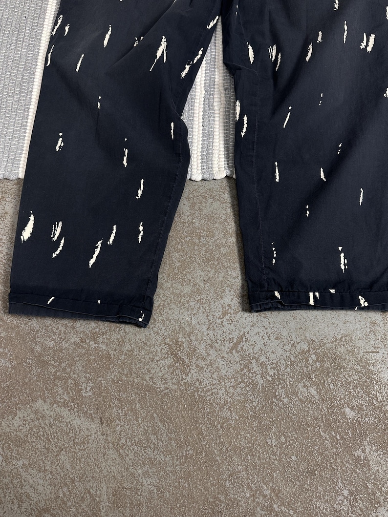 Puede incluir: Pantalones azul marino oscuro con un patr&oacute;n abstracto blanco de pinceladas. Los pantalones est&aacute;n doblados en el dobladillo. El fondo incluye una alfombra a rayas grises y blancas y una superficie de hormig&oacute;n gris.