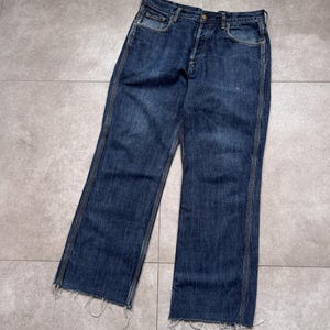 Puede incluir: Vaqueros de mezclilla azul oscuro con bragueta de botones y dobladillo deshilachado. Los vaqueros tienen un dise&ntilde;o cl&aacute;sico de cinco bolsillos y un corte de pierna recta. La mezclilla tiene un aspecto desgastado.