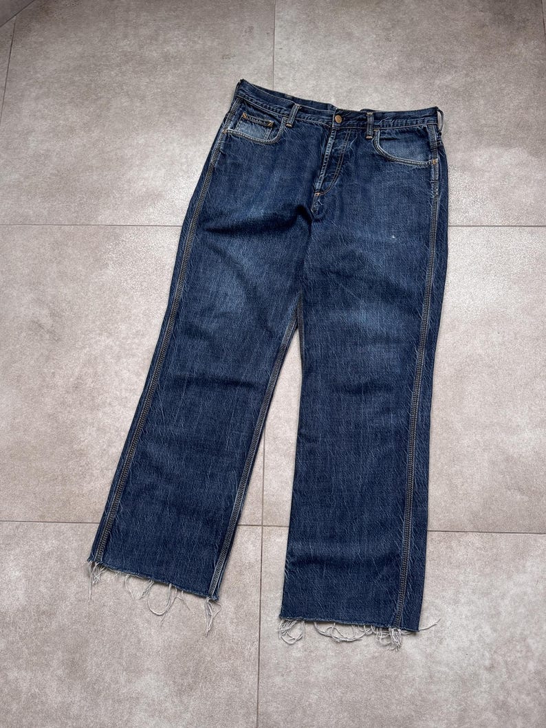 Puede incluir: Vaqueros de mezclilla azul oscuro con bragueta de botones y dobladillo deshilachado. Los vaqueros tienen un dise&ntilde;o cl&aacute;sico de cinco bolsillos y un corte de pierna recta. La mezclilla tiene un aspecto desgastado.