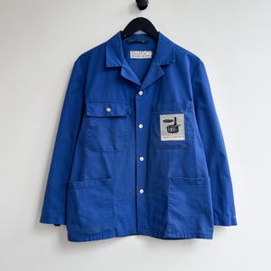 Può includere: Una giacca da lavoro blu con una toppa sul petto sinistro che dice "WORKSHOP" e ha un'immagine di un edificio. La giacca ha due grandi tasche sul davanti.