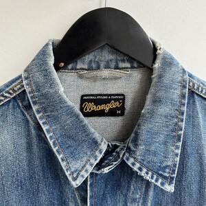May include: A light blue denim jacket with a Wrangler label. The label says "Original Styling & Features" and has the Wrangler logo in gold lettering. The jacket is size M.