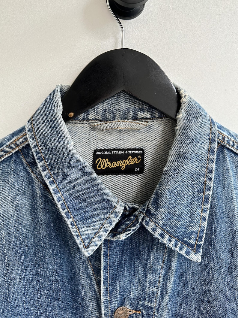 May include: A light blue denim jacket with a Wrangler label. The label says "Original Styling & Features" and has the Wrangler logo in gold lettering. The jacket is size M.