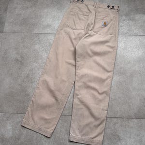 Puede incluir: Pantalones de trabajo color beige con dise&ntilde;o de pierna recta. Los pantalones tienen dos bolsillos traseros y un peque&ntilde;o parche con el logotipo. La cintura tiene cierres de bot&oacute;n. Los pantalones est&aacute;n hechos de una tela duradera.