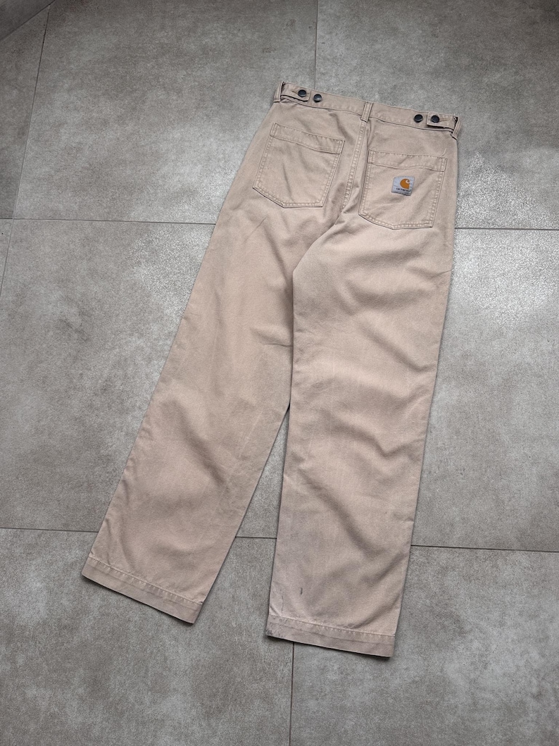 Puede incluir: Pantalones de trabajo color beige con dise&ntilde;o de pierna recta. Los pantalones tienen dos bolsillos traseros y un peque&ntilde;o parche con el logotipo. La cintura tiene cierres de bot&oacute;n. Los pantalones est&aacute;n hechos de una tela duradera.