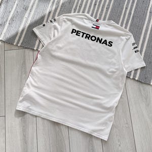 May include: White t-shirt with the word "PETRONAS" printed on the back in black. The shirt has a red and white stripe on the left sleeve and a small Tommy Hilfiger logo on the back of the collar.
