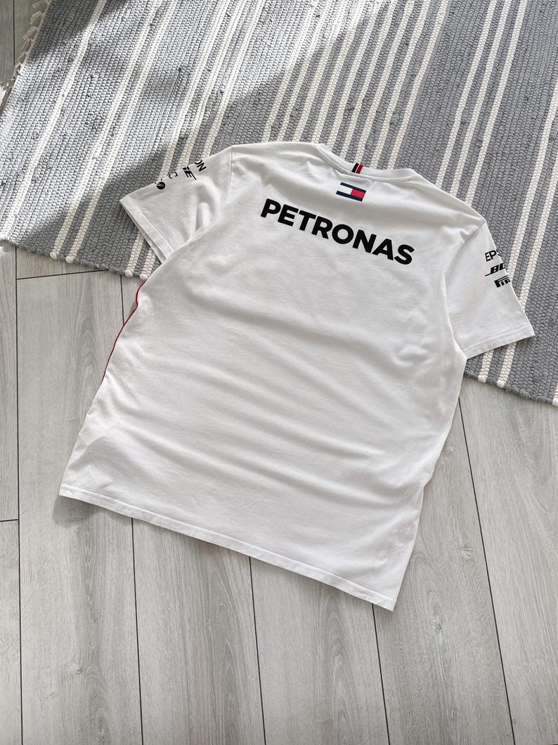 May include: White t-shirt with the word "PETRONAS" printed on the back in black. The shirt has a red and white stripe on the left sleeve and a small Tommy Hilfiger logo on the back of the collar.