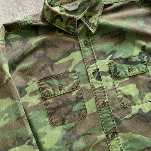 May include: A green, brown, and black camouflage long-sleeve button-up shirt with two chest pockets. The shirt has a label that reads "Five Brother" on the collar.