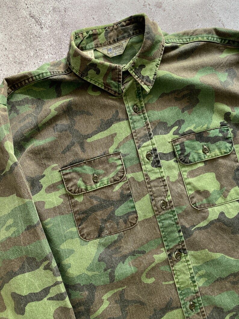 May include: A green, brown, and black camouflage long-sleeve button-up shirt with two chest pockets. The shirt has a label that reads "Five Brother" on the collar.