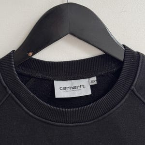May include: Black crew neck sweatshirt with the Carhartt Work In Progress logo on a white tag. The sweatshirt is hanging on a black hanger. The tag also indicates the size is XS.