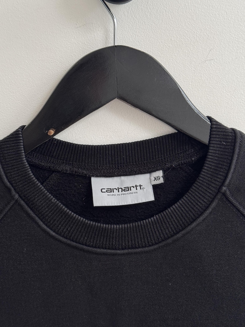 May include: Black crew neck sweatshirt with the Carhartt Work In Progress logo on a white tag. The sweatshirt is hanging on a black hanger. The tag also indicates the size is XS.