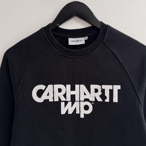 May include: Black sweatshirt with the white text "CARHARTT WIP" across the chest. The sweatshirt has a crew neck, raglan sleeves, and a kangaroo pocket. The Carhartt logo is on the neck label.