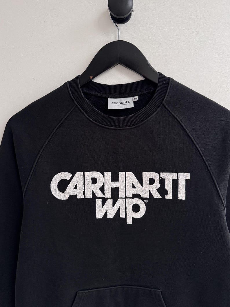 May include: Black sweatshirt with the white text "CARHARTT WIP" across the chest. The sweatshirt has a crew neck, raglan sleeves, and a kangaroo pocket. The Carhartt logo is on the neck label.
