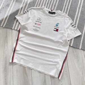 May include: White t-shirt with the Mercedes-AMG Petronas Formula One Team logo, sponsors' logos, and the Tommy Hilfiger logo. The shirt has red and blue stripes on the sides.