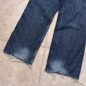 Puede incluir: Primer plano de unos vaqueros de mezclilla azules con un dobladillo deshilachado y desgastado. Los vaqueros tienen un lavado descolorido y un corte relajado. La tela muestra signos de desgaste, lo que a&ntilde;ade a la est&eacute;tica vintage. Los vaqueros est&aacute;n planos sobre una superficie embaldosada.