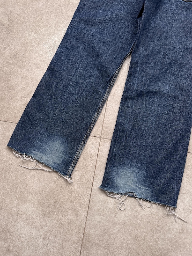 Puede incluir: Primer plano de unos vaqueros de mezclilla azules con un dobladillo deshilachado y desgastado. Los vaqueros tienen un lavado descolorido y un corte relajado. La tela muestra signos de desgaste, lo que a&ntilde;ade a la est&eacute;tica vintage. Los vaqueros est&aacute;n planos sobre una superficie embaldosada.