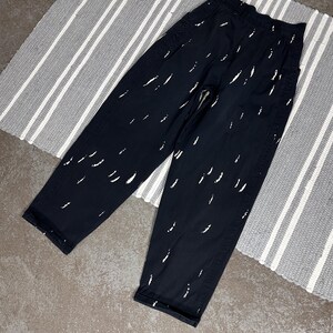 Puede incluir: Pantalones negros con un patr&oacute;n de pinceladas blancas. Los pantalones tienen un corte holgado y una cintura alta. Los pantalones est&aacute;n sobre una alfombra a rayas grises y blancas.