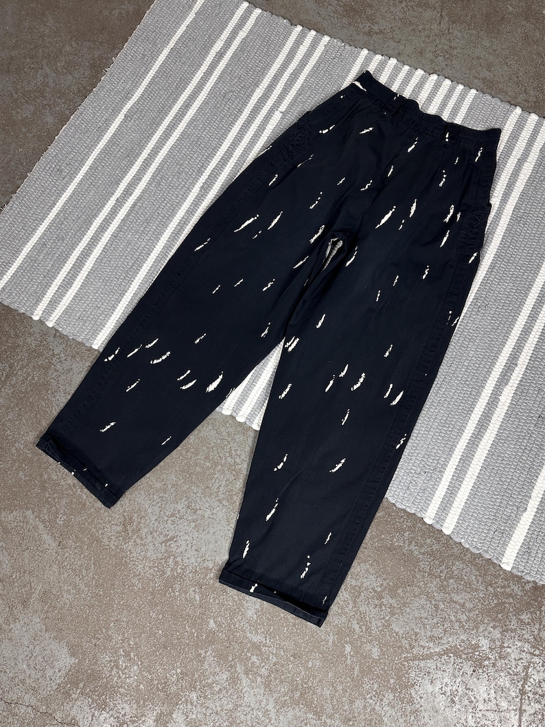 Puede incluir: Pantalones negros con un patr&oacute;n de pinceladas blancas. Los pantalones tienen un corte holgado y una cintura alta. Los pantalones est&aacute;n sobre una alfombra a rayas grises y blancas.