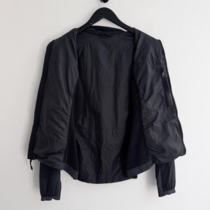 May include: Black jacket hanging on a black hanger against a white wall. The jacket is open, revealing the interior lining and a zippered pocket. The sleeves are long and the jacket appears to be made of a lightweight material.