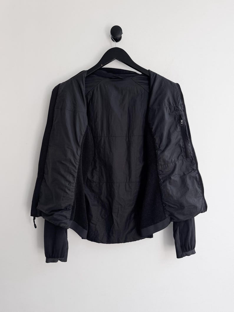 May include: Black jacket hanging on a black hanger against a white wall. The jacket is open, revealing the interior lining and a zippered pocket. The sleeves are long and the jacket appears to be made of a lightweight material.