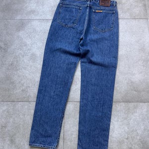 Pode incluir: Jeans azuis de ganga com um design cl&aacute;ssico de cinco bolsos. Os jeans apresentam um corte de perna reto e um patch de couro castanho com o nome da marca nas costas. Os jeans est&atilde;o estendidos numa superf&iacute;cie cinzenta.